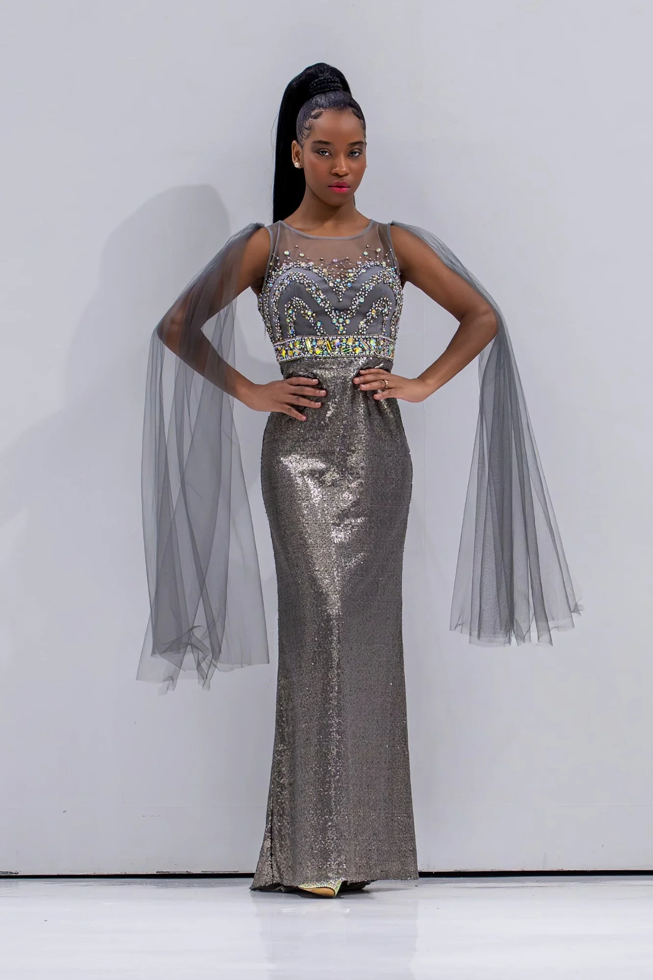 A woman in a glamorous silver gown with sequins and beading, standing confidently with her hands on her hips against a plain white background.