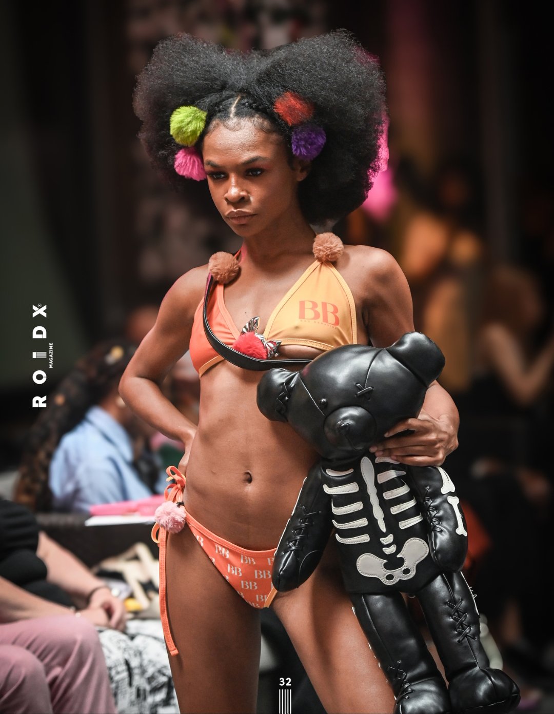 Model walking runway wearing a bikini with pom-poms and holding a teddy bear-shaped purse, both in black leather with skeleton print, on a fashion show runway.