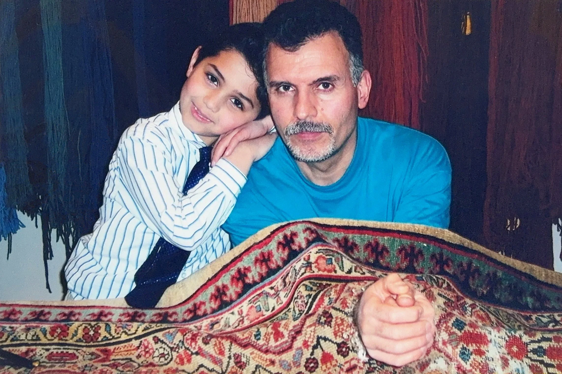 A young boy with dark hair leaning his head on an older man's shoulder, both smiling slightly. They are sitting behind a colorful patterned table or cloth, with a dark wooden background.