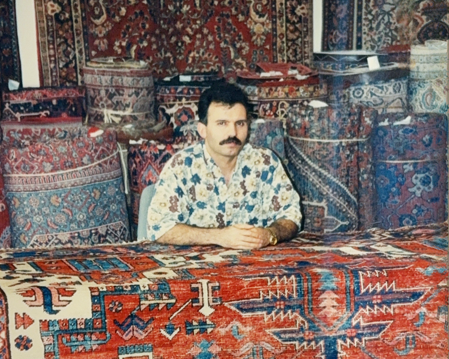 A man with a mustache, wearing a floral patterned shirt, sitting at a table covered with a red and blue Persian rug design, with a background of stacked Persian rugs hanging on the wall.