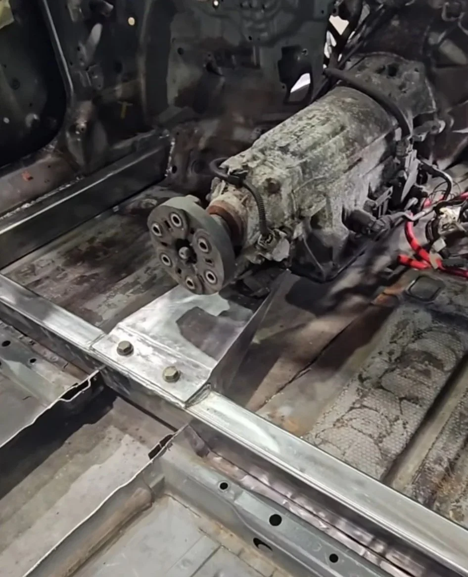 Close-up of a car's stripped interior showing an exposed engine or transmission mounted on a metal frame, with nearby wiring and metal panels, indicating vehicle repair or modification.