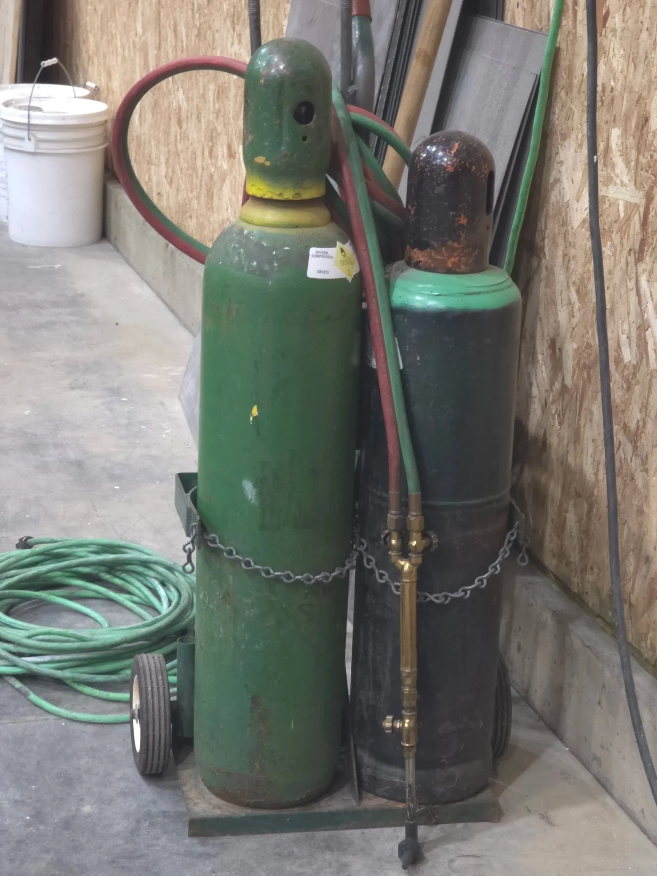 Two old welding gas cylinders, one green and one black, with hoses draped over them, chained together in a workshop setting.
