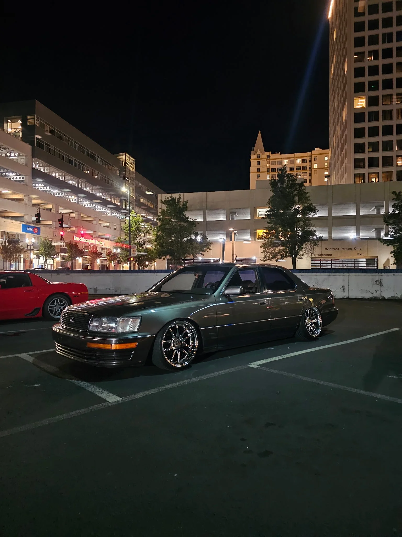 A nighttime parking lot scene with a vintage silver car with chrome wheels in foreground and a red sports car in the background. City buildings with lights and trees are visible behind the cars.