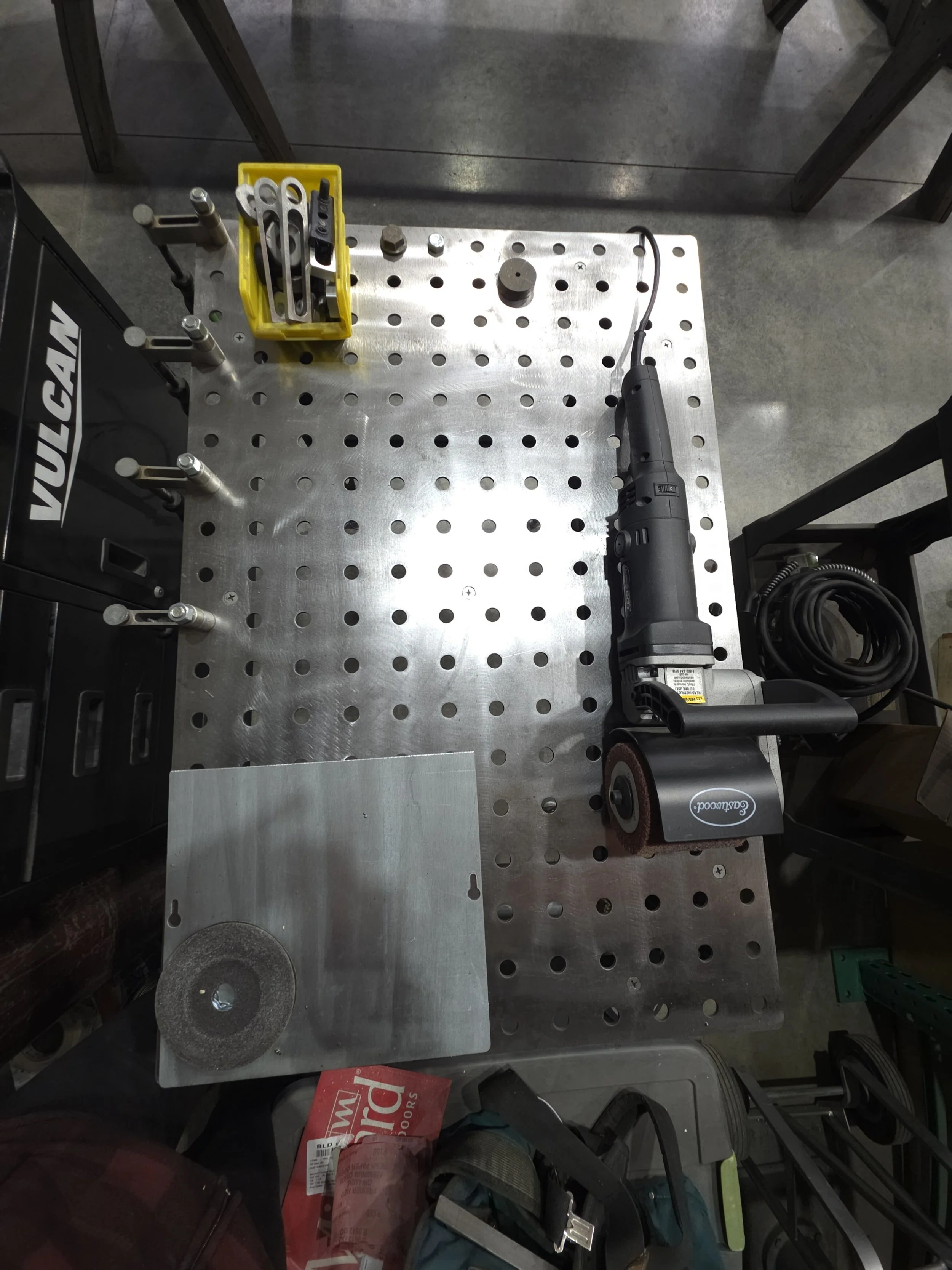 Workstation with a metal pegboard table, a cordless angle grinder, a yellow toolbox with metal tools, a metal square, and some miscellaneous tools and objects.