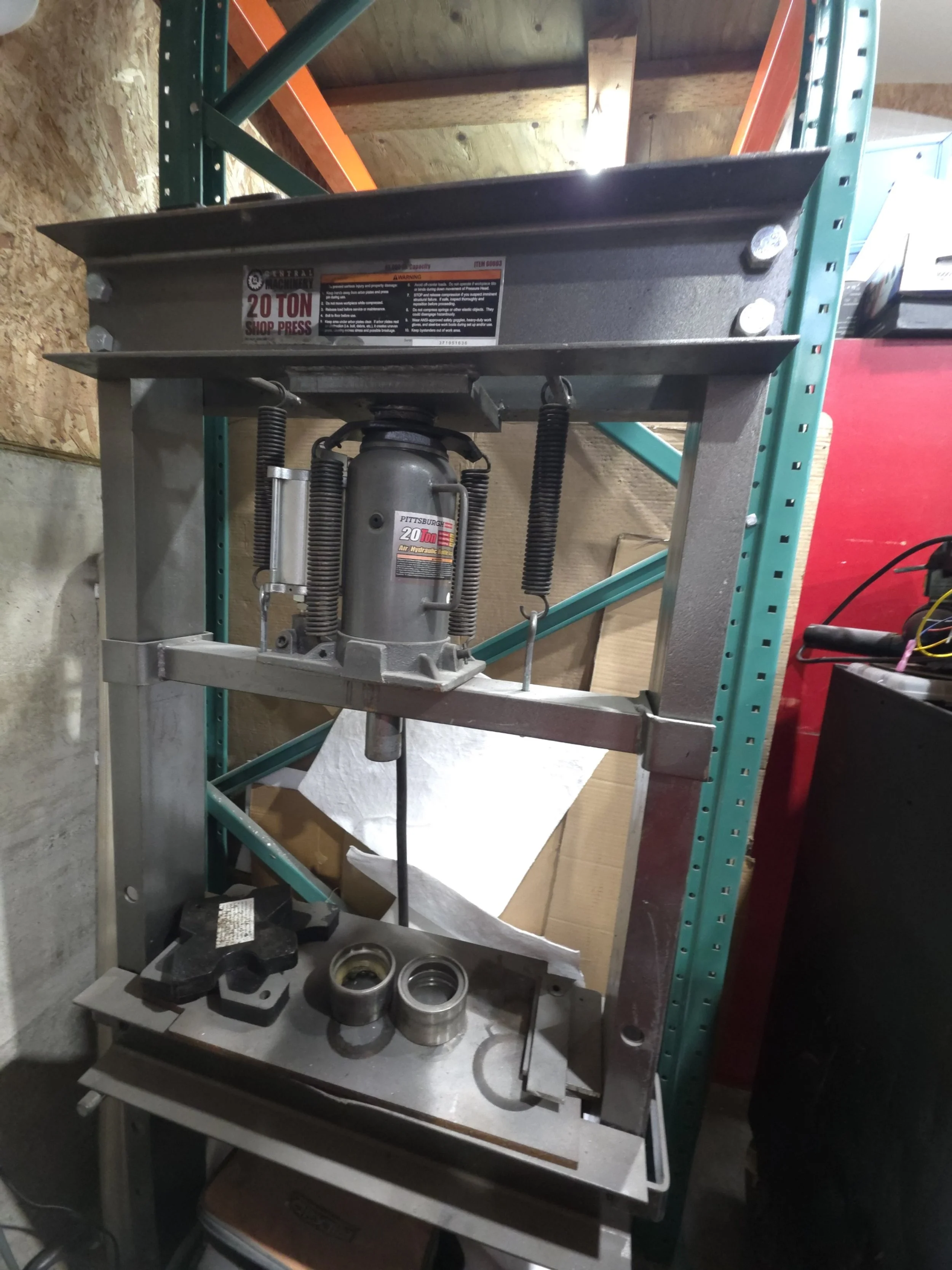 A 20-ton shop press on a metal stand with various metal parts and bearings on a lower shelf, located in a workshop or garage.