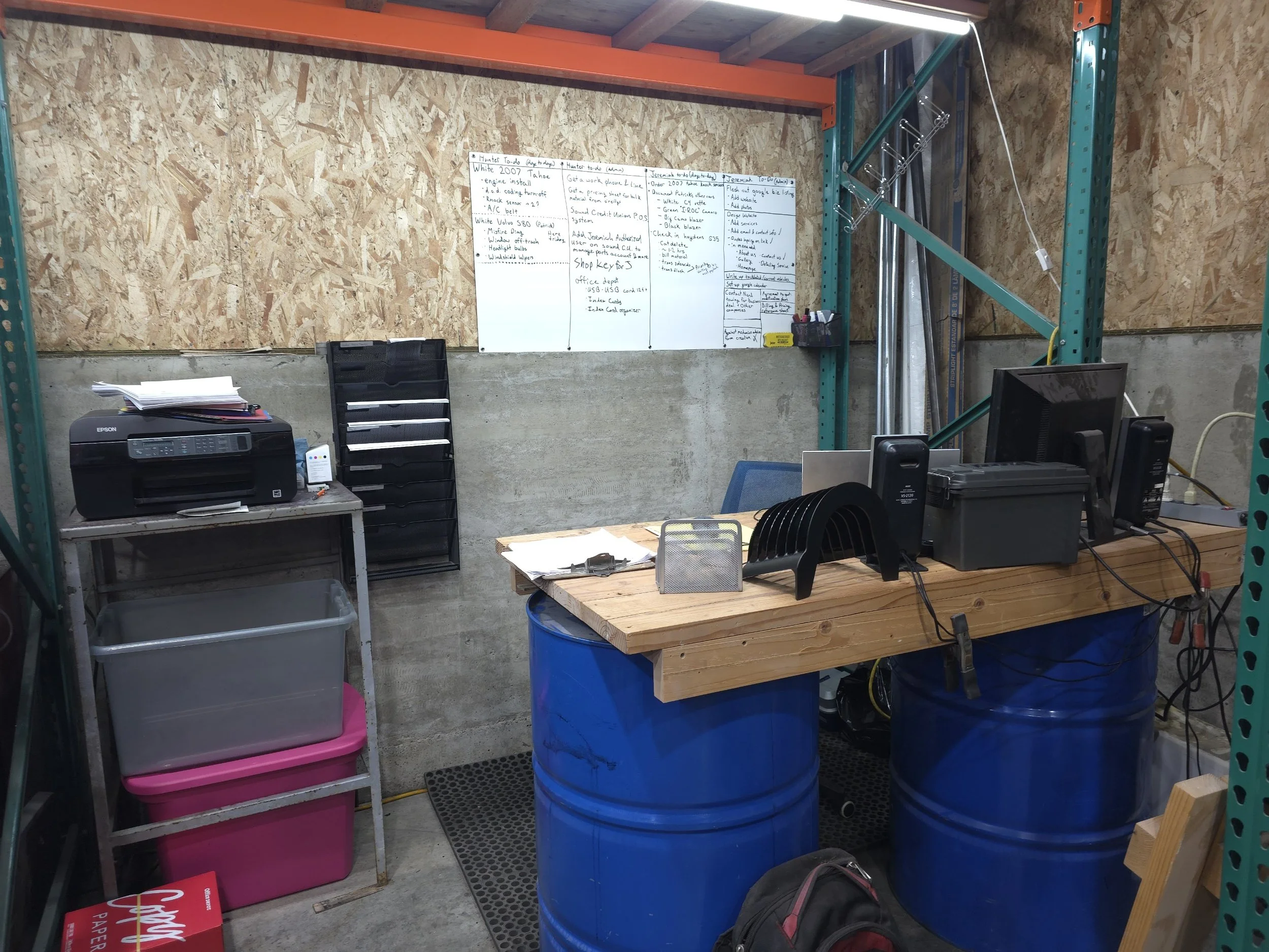 A cluttered workspace with a wooden desk supported by two blue oil drums, holding computer monitors, a printer, and office supplies. Behind the desk, walls are partially concrete and plywood, with a whiteboard filled with notes and a small black storage rack. To the left, a pink storage bin and a black document holder are on a silver table, with a printer and various papers stacked on top. A black backpack is on the floor.