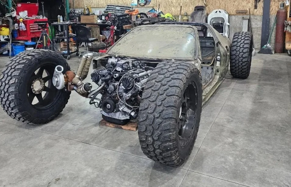 A partially assembled off-road vehicle or dune buggy in a workshop, with large rugged tires, exposed engine, and no body panels.