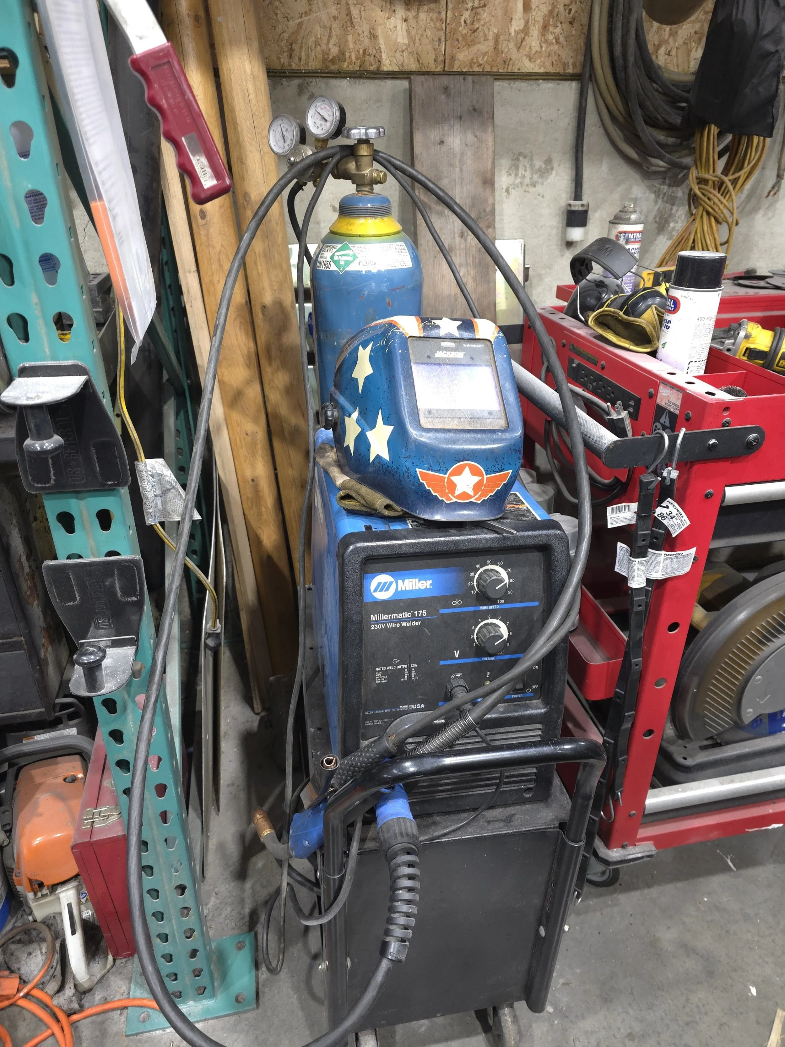 A welding station with a Miller Millermatic 175 wire welder, a welding helmet with a patriotic stars and stripes design, and a propane tank, in a workshop setting.
