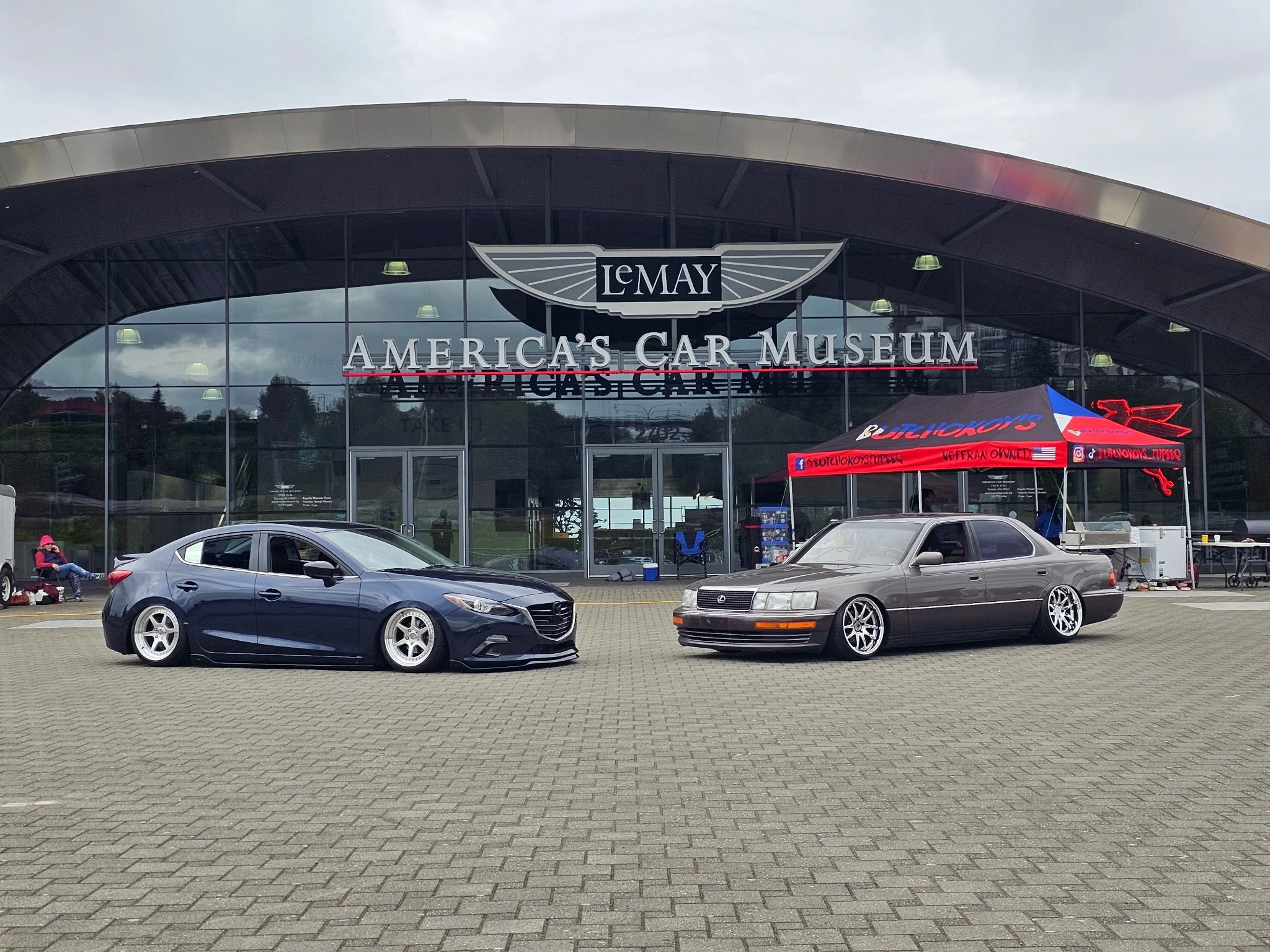 Two cars parked in front of the LeMay America’s Car Museum with a building in the background.