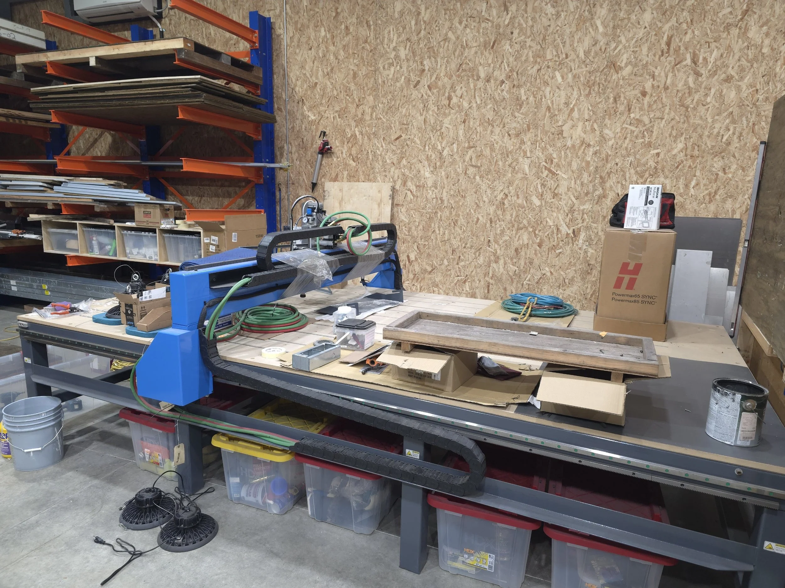 Workshop with a large work table holding tools, wires, boxes, a saw, and a wood frame. Shelves with materials and equipment are in the background.