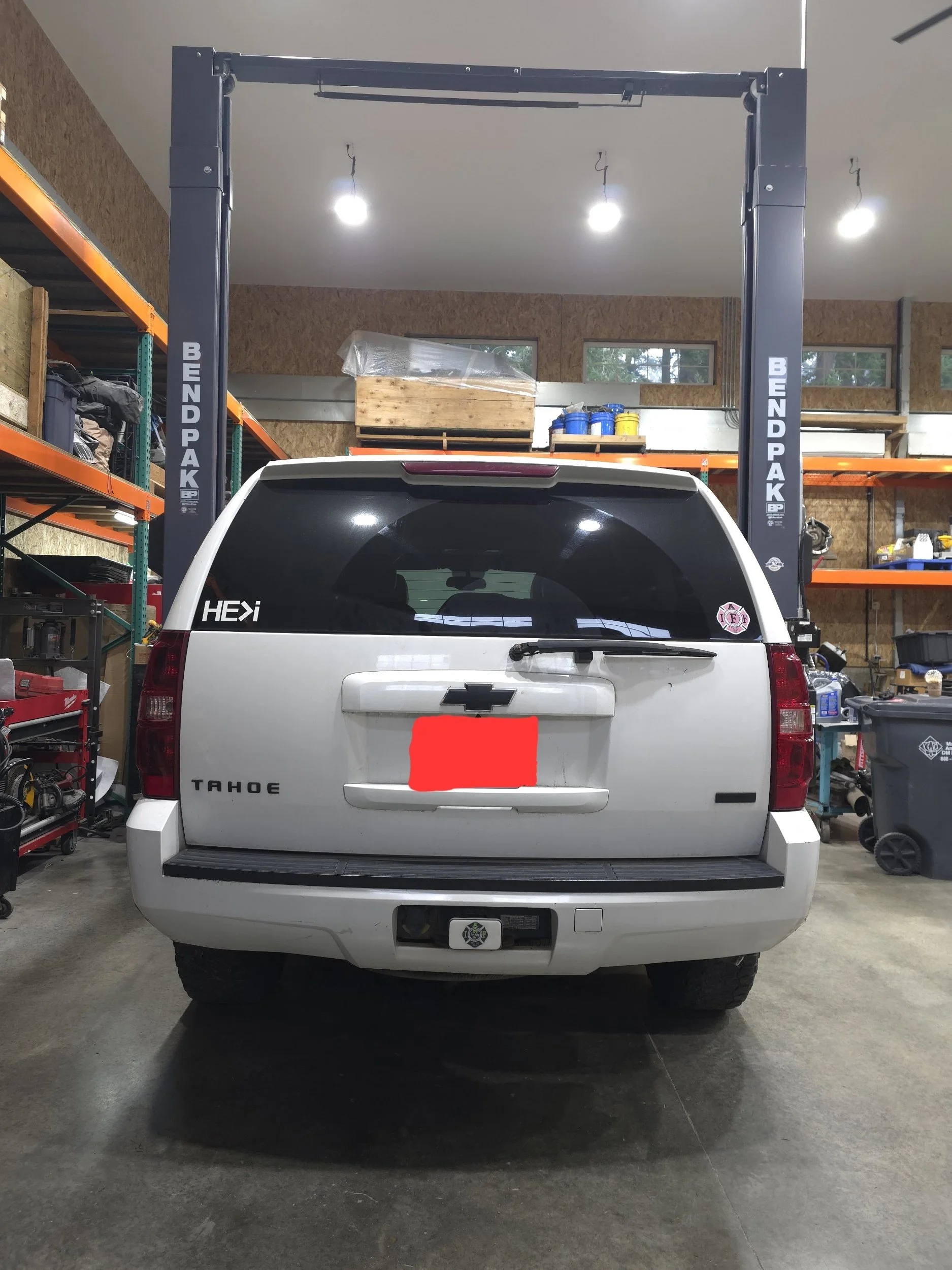 Rear view of a white Chevrolet Tahoe inside a garage or workshop with shelving and tools, under a vehicle lift.