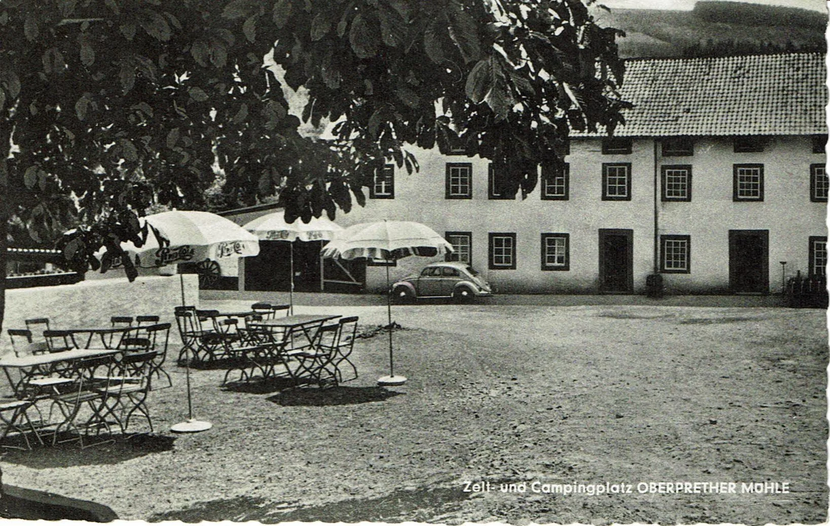Schwarz-Weiß-Foto eines Campingplatzes mit Tischen, Stühlen, Sonnenschirmen, einem Gebäude im Hintergrund, einem Auto und Bäumen, mit der Beschriftung "Zelt- und Campingplatz Oberprether Mühle".