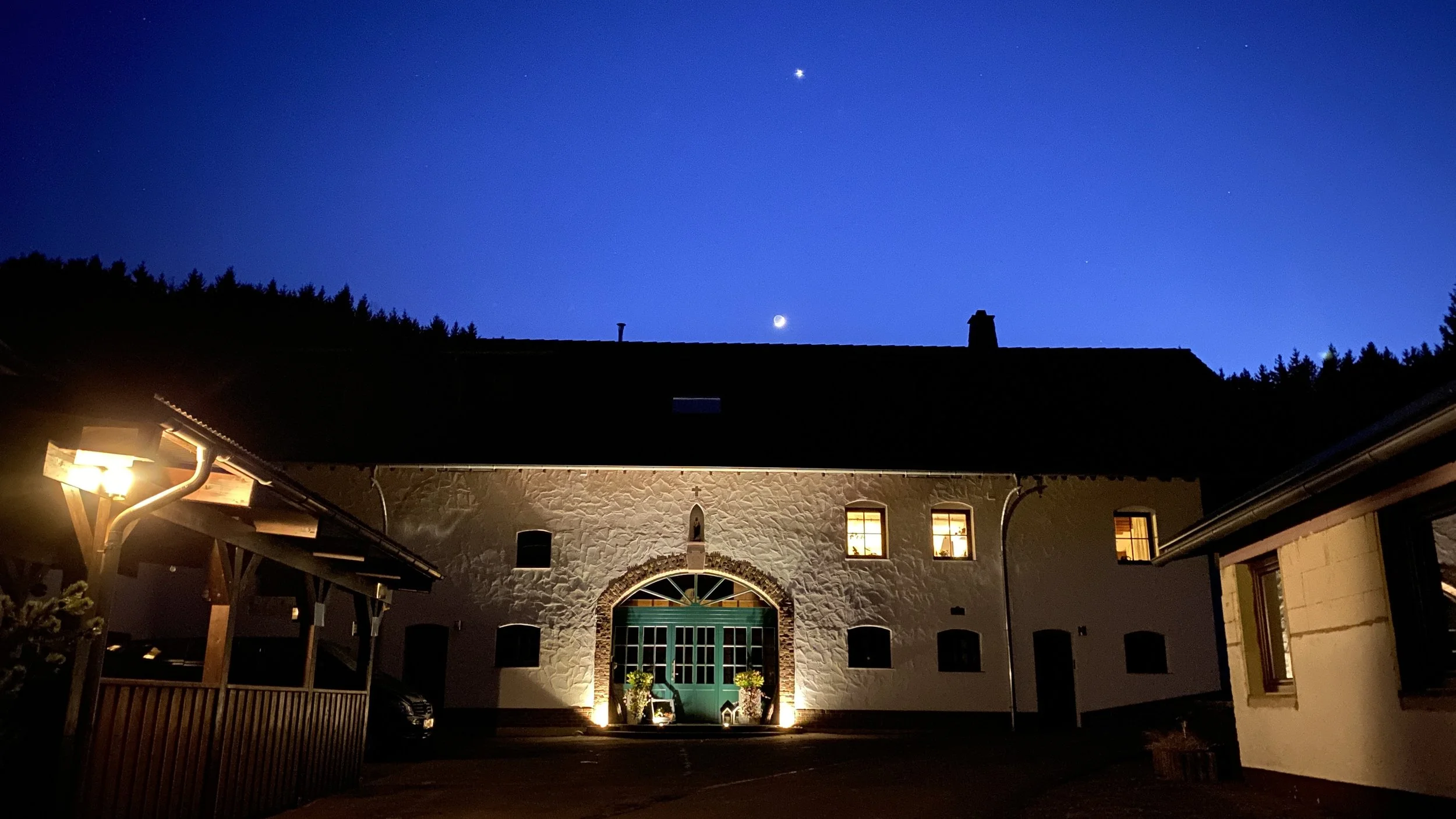 Ein Haus bei Nacht mit beleuchteter Eingangstür, Fenstern und außenstehender Beleuchtung. Über dem Haus ist der Himmel mit sichtbarem Mond, Venus und einigen Sternen.