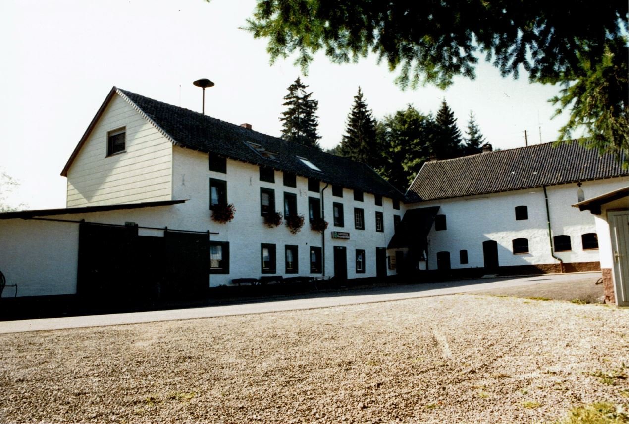Ein weißes, mehrstöckiges Bauernhaus mit dunklen Fenstern und Blumenampeln vor einem Hintergrund von Tannenbäumen.