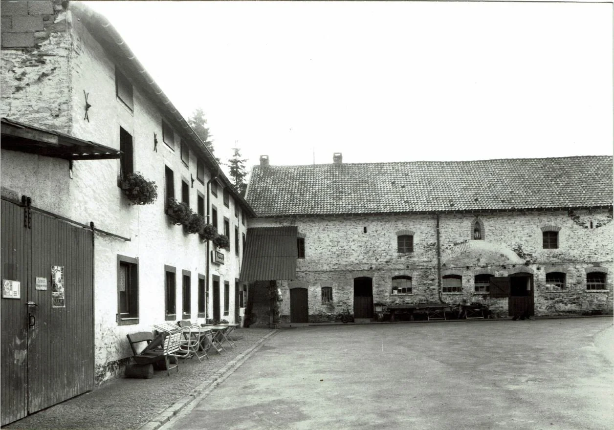 Schwarz-weiß-Foto von einem alten Hof mit gemauertem Gebäude, Holztüren, Fenstern mit Blumentöpfen, Tischen und Bänken im Innenhof.