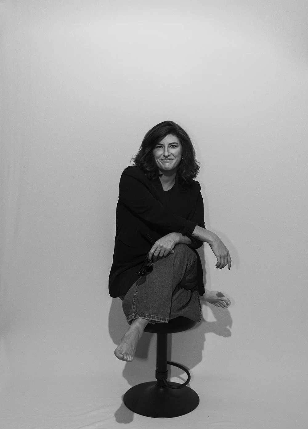 A woman sitting on a stool with a plain white background, wearing a black top and jeans, smiling slightly.