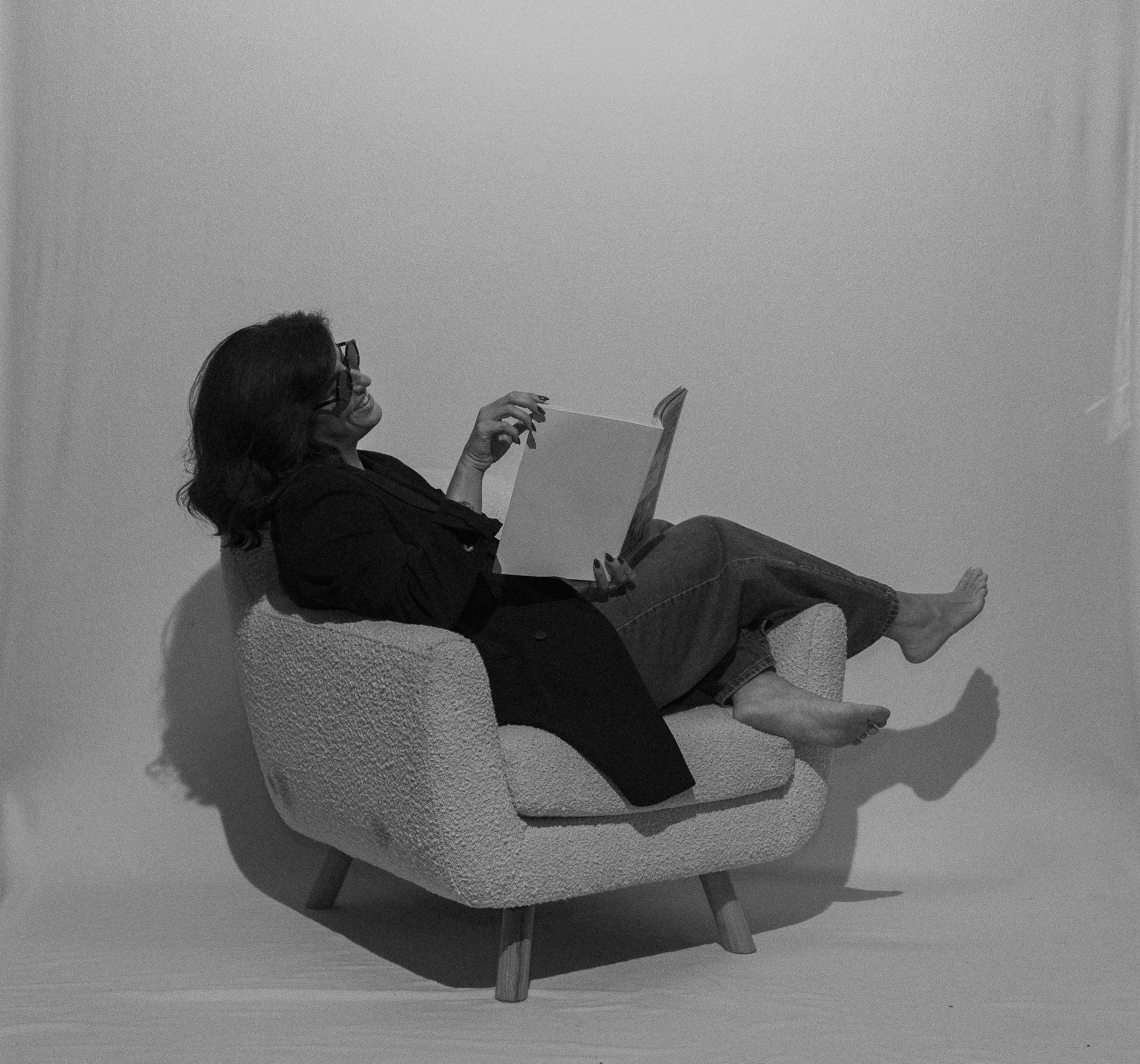 A woman with glasses is reclining on a cozy, textured armchair, reading a book with a relaxed and cheerful demeanor.