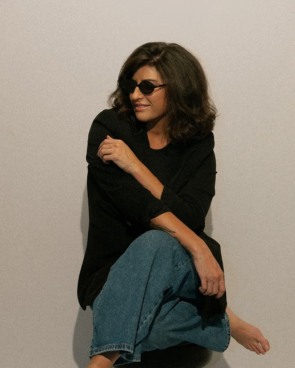 A woman with shoulder-length wavy brown hair wearing black sunglasses, a black top, and blue jeans, sitting against a plain white wall, smiling and looking to the side.