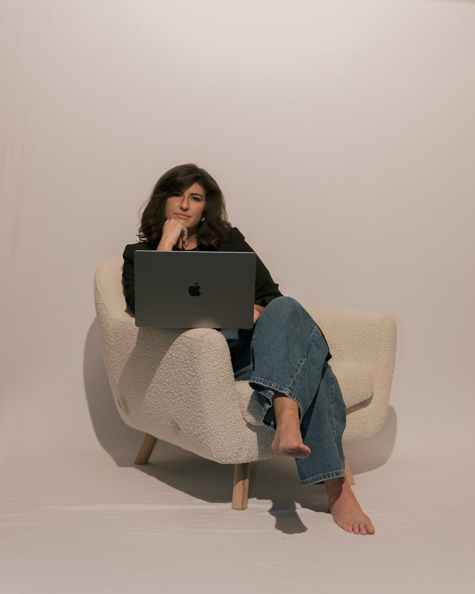 Woman sitting on a cream-colored armchair with wooden legs, using a silver Apple MacBook. She has shoulder-length dark hair, is wearing a black blazer and blue jeans, with one barefoot and the other foot resting on the chair. Her pose is relaxed, and she is resting her chin on her hand, looking directly at the camera against a plain white background.