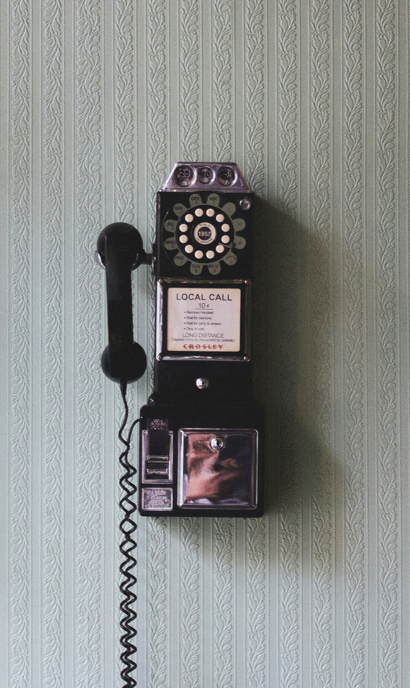 A vintage black rotary payphone mounted on a patterned green wall.