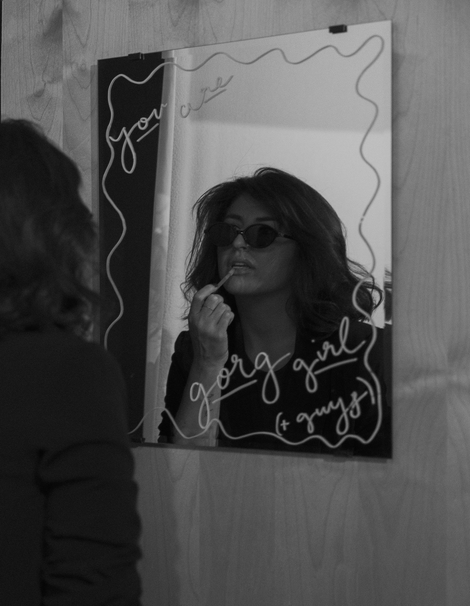 Woman with wavy hair wearing sunglasses, looking at her reflection in a mirror, with handwritten text on the mirror that says 'you are special' or similar, and her lips are slightly parted.