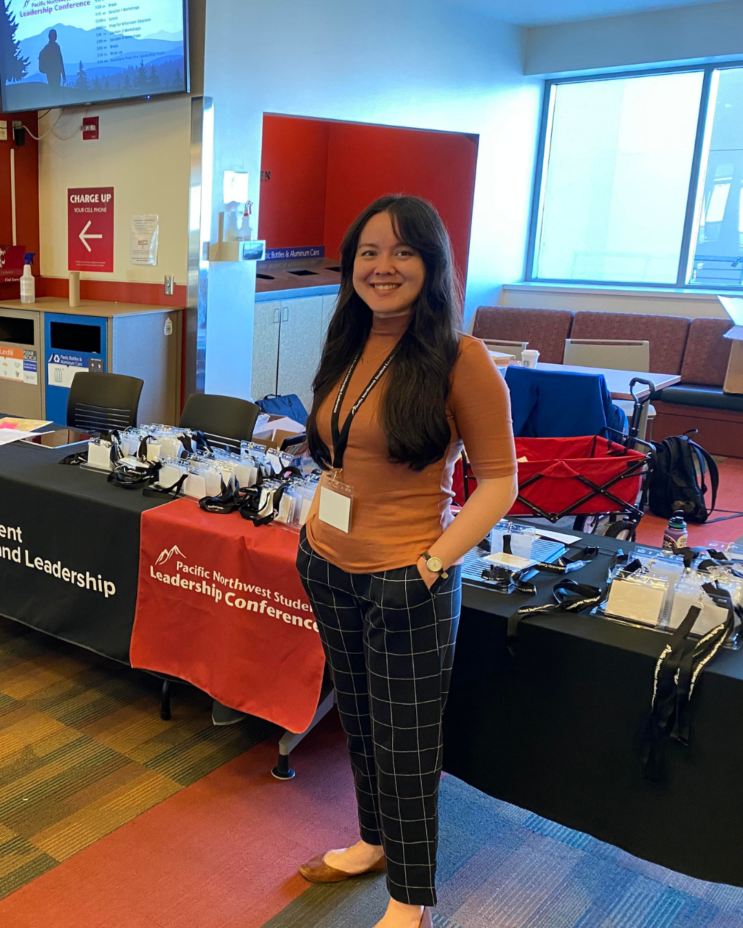A woman with long dark hair smiling at a conference, standing next to a table with conference badges, lanyards, and promotional materials. She is wearing a brown top, checkered pants, and a watch, in a room with large windows and a red and black conference banner.