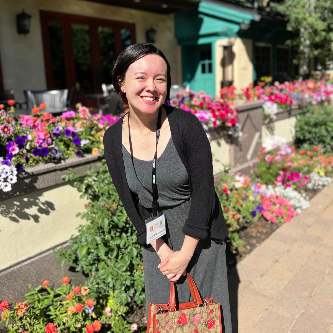 A woman with short dark hair smiling outdoors on a sunny day, holding a strawberry-patterned tote bag, with a colorful flower bed and a building with a teal awning in the background.