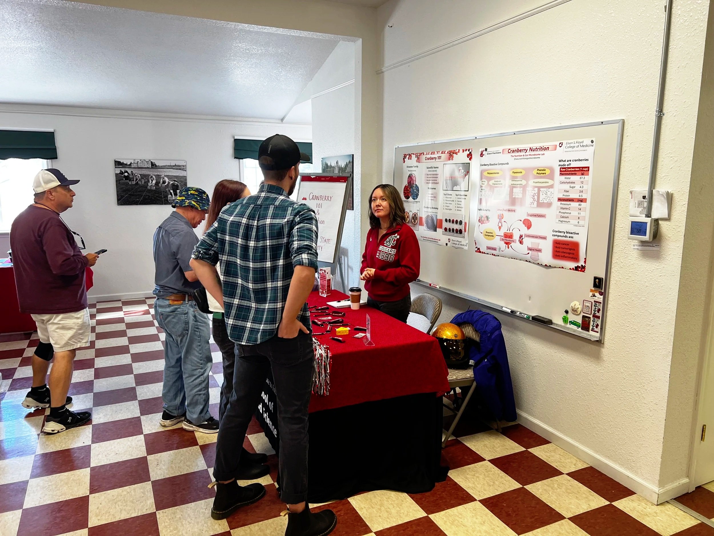 People at a science or health fair booth discussing cranberry research, with informational posters on the wall and items on the table, inside a room with checkered flooring and white walls.