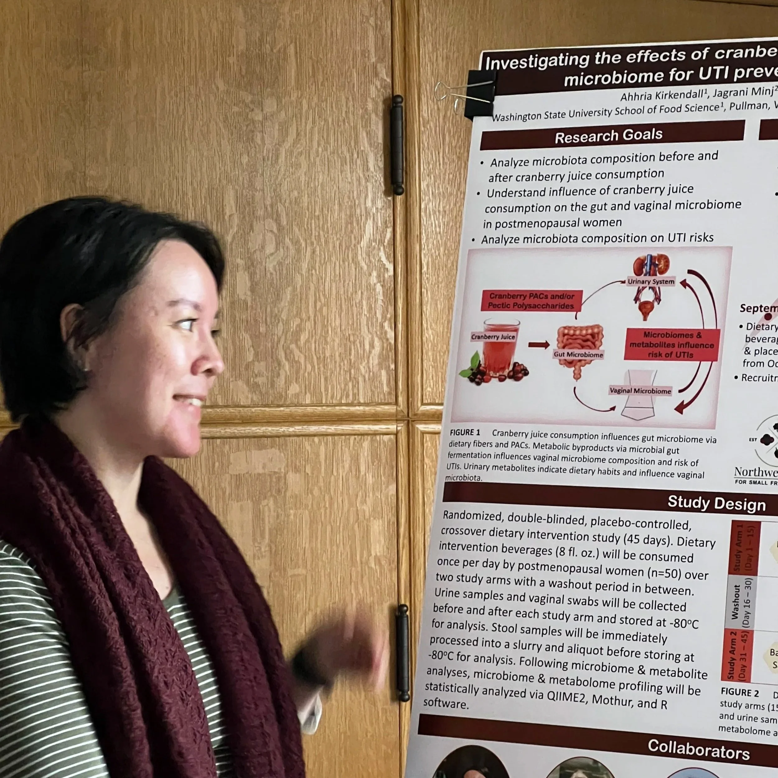 A woman standing next to a scientific research poster on a wood-paneled wall, speaking or gesturing with her hand, with detailed text and diagrams discussing cranberry juice, gut microbiome, and study design.