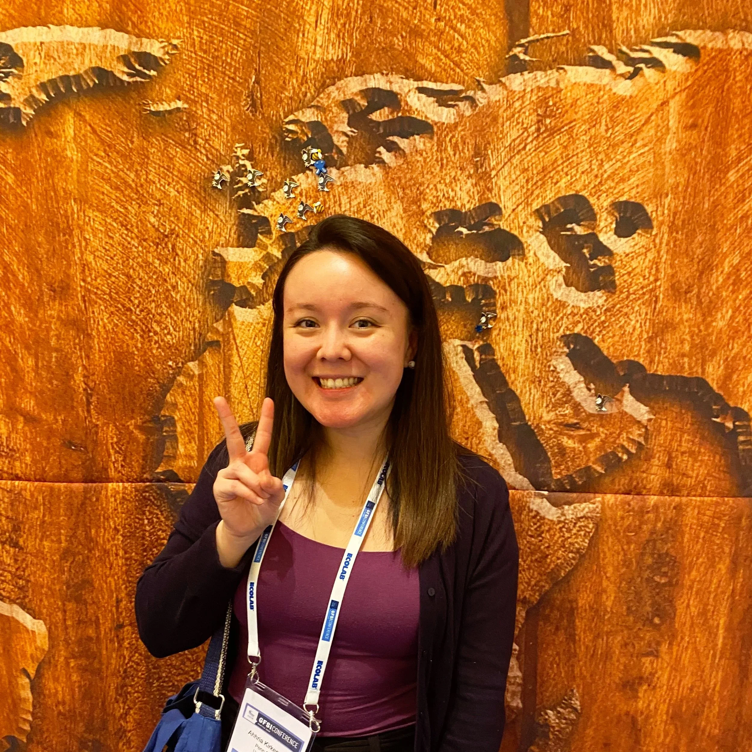 A smiling woman making a peace sign in front of a large rock wall with a carved paw print.