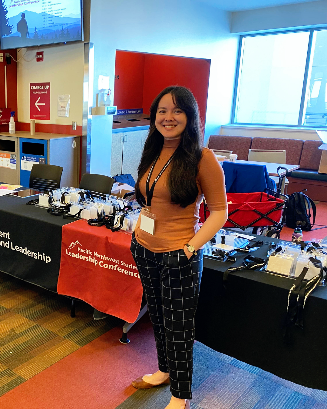 A smiling woman with long dark hair stands at a registration table for the Pacific Northwest Student Leadership Conference. The table has conference badges, lanyards, and name tags, with a black tablecloth and a red cover. In the background, there's a large window, red wall, and a screen showing the conference schedule.