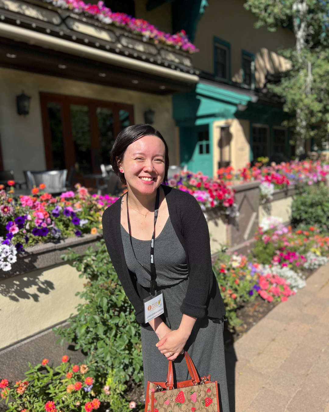 A woman, The Cranberry Doctor, with black hair smiling outdoors on a sunny day, standing in front of a building with colorful flower boxes on the windowsill, wearing a gray dress, black cardigan, and holding a bag with strawberry print.