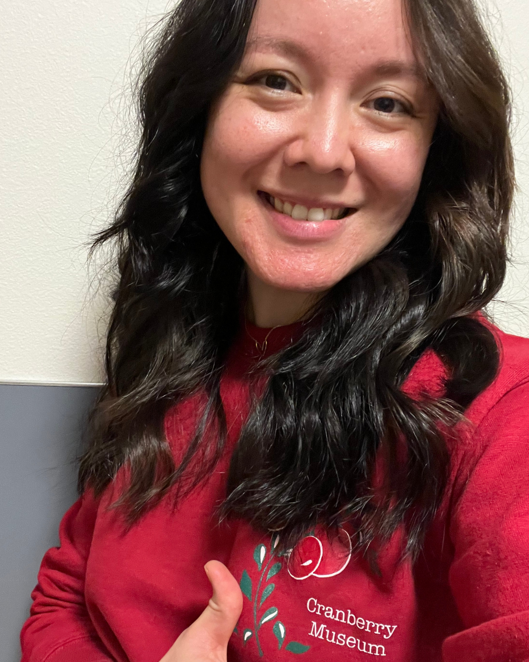 A woman, The Cranberry Doctor, with long dark wavy hair, smiling and showing her teeth, wearing a red sweatshirt with a logo and text that reads 'Cranberry Museum,' giving a thumbs-up.