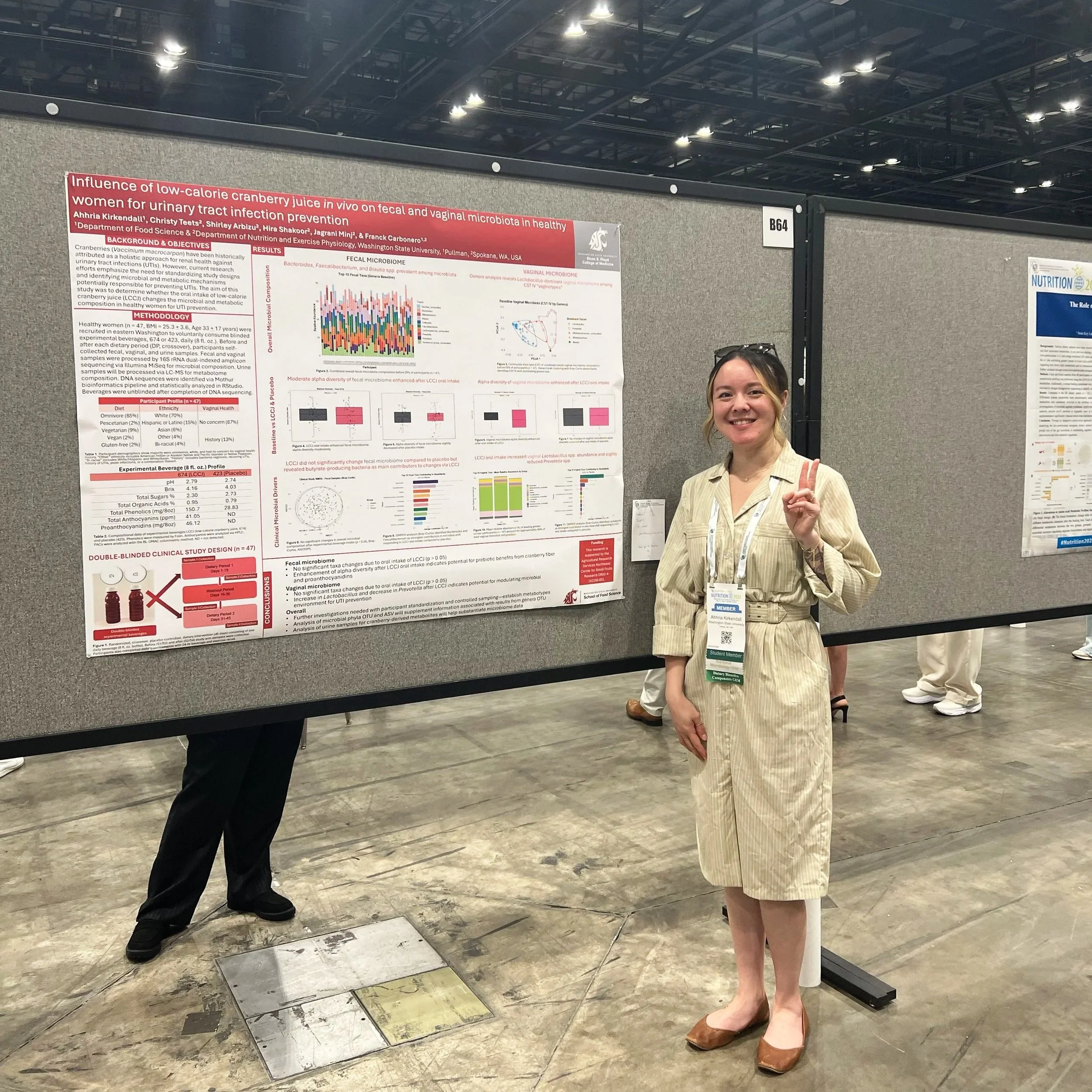 A woman standing next to a research poster at a conference, making a peace sign and smiling.