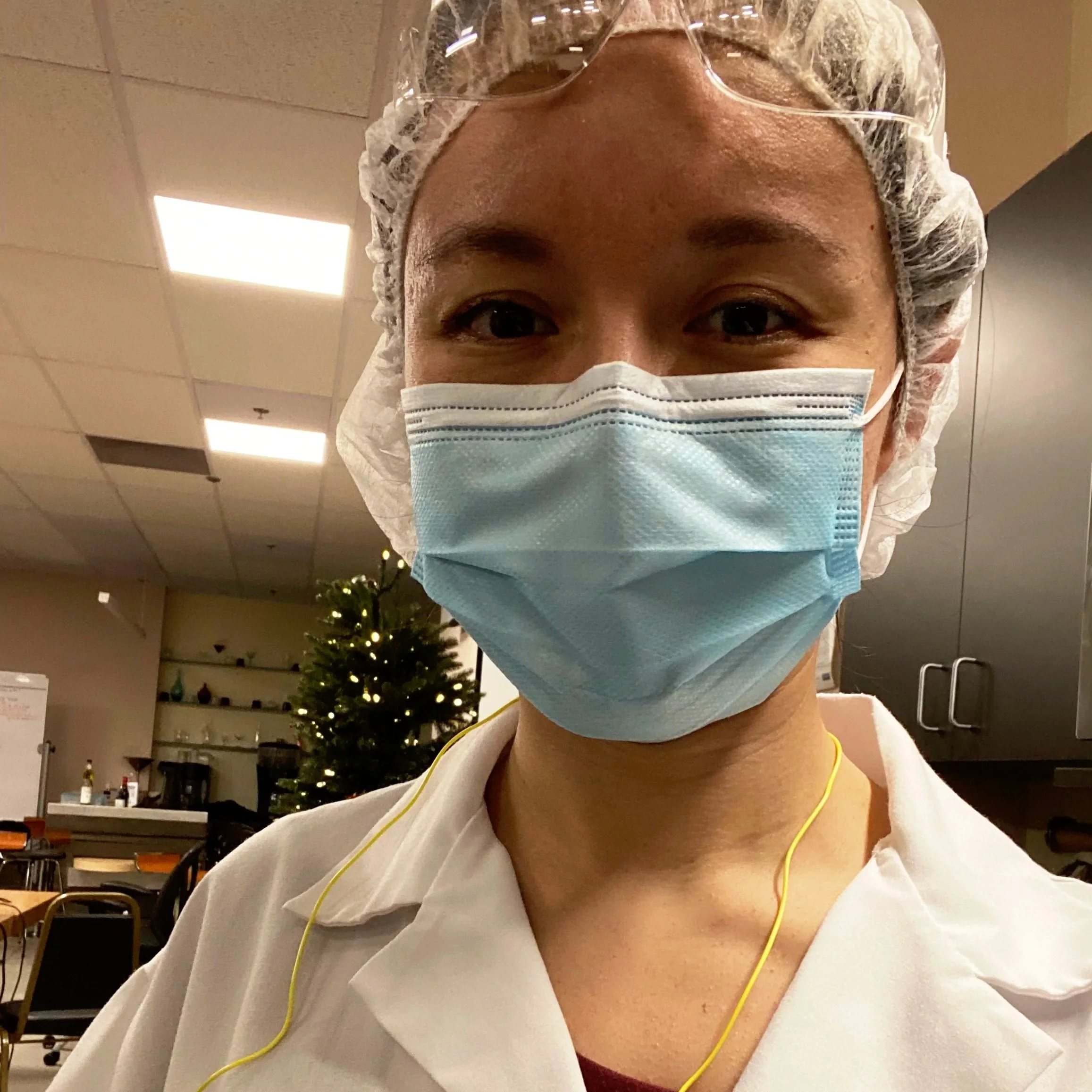 A healthcare worker wearing a white uniform, a blue face mask, protective goggles, and a hairnet, standing in a room with a Christmas tree in the background.