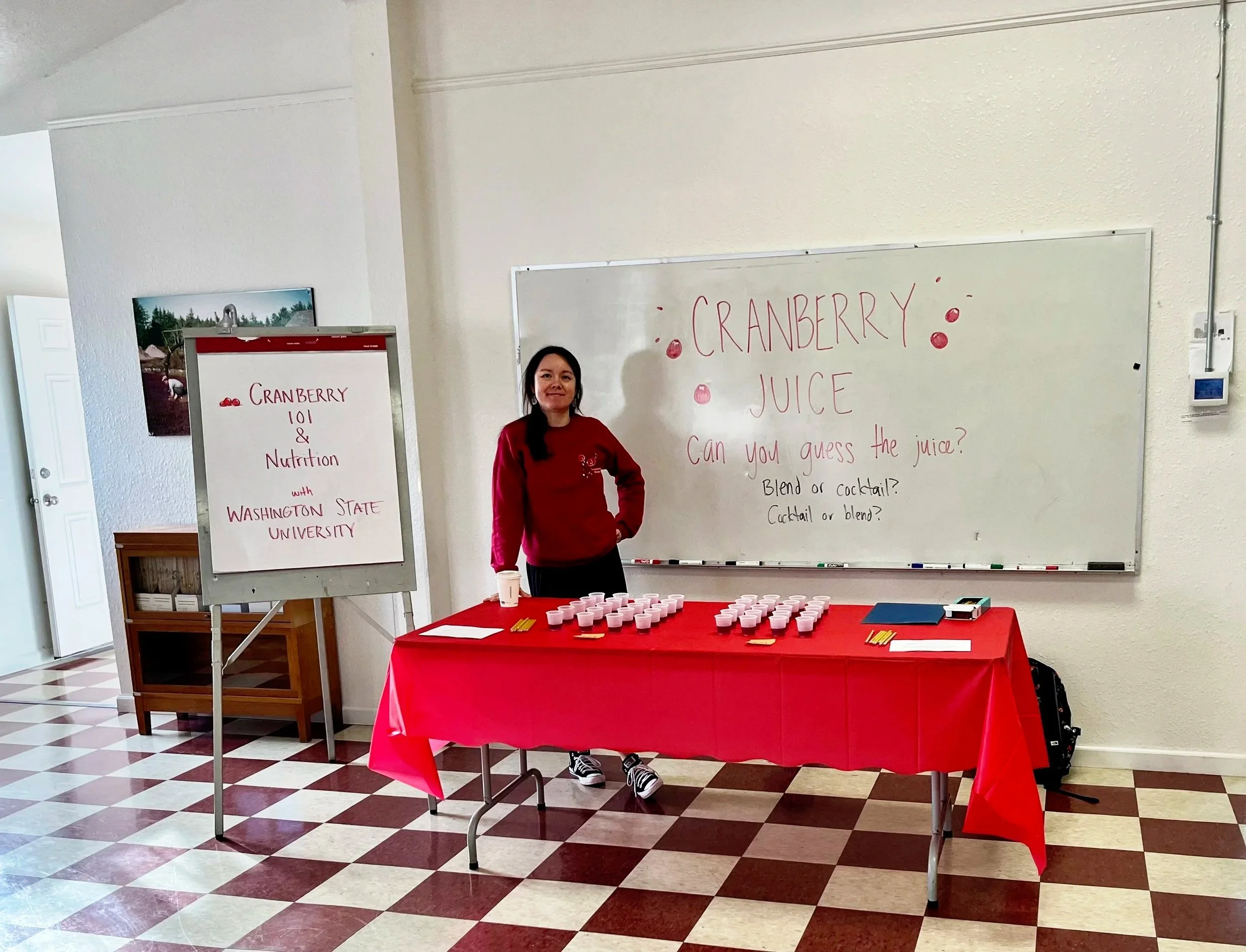 A woman stands behind a table with cups of cranberry juice, set up for a presentation or tasting event about cranberry juice, with a whiteboard and an easel displaying information about cranberry nutrition and Washington State University.
