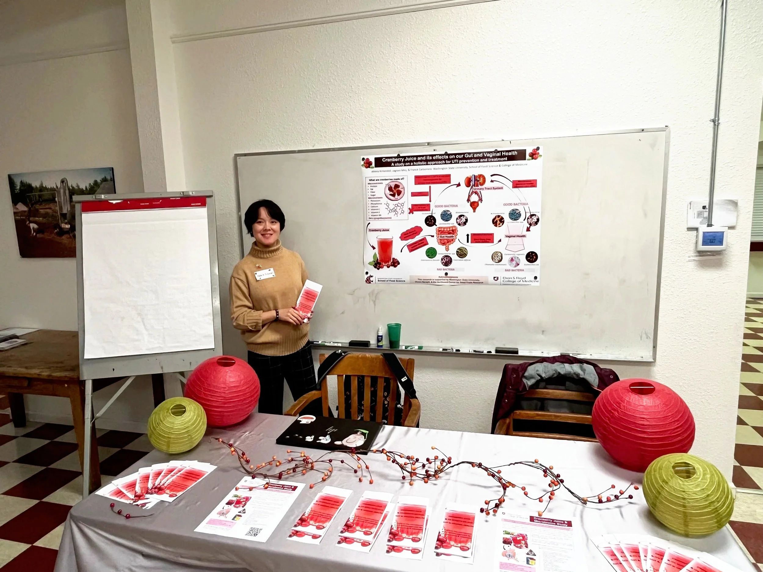 A woman standing at a table with informational flyers, red and yellow decorative lanterns, and decorations, in front of a whiteboard displaying a health-related poster about cranberries and gut health. The woman is smiling and holding a pamphlet, with a name tag on her tan sweater.