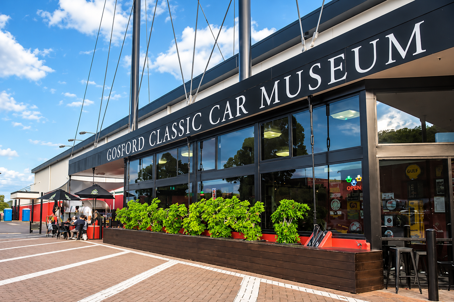Exterior of Gosford Classic Car Museum with large glass windows, green plants in a planter, outdoor seating area, and people dining under umbrellas on a sunny day.