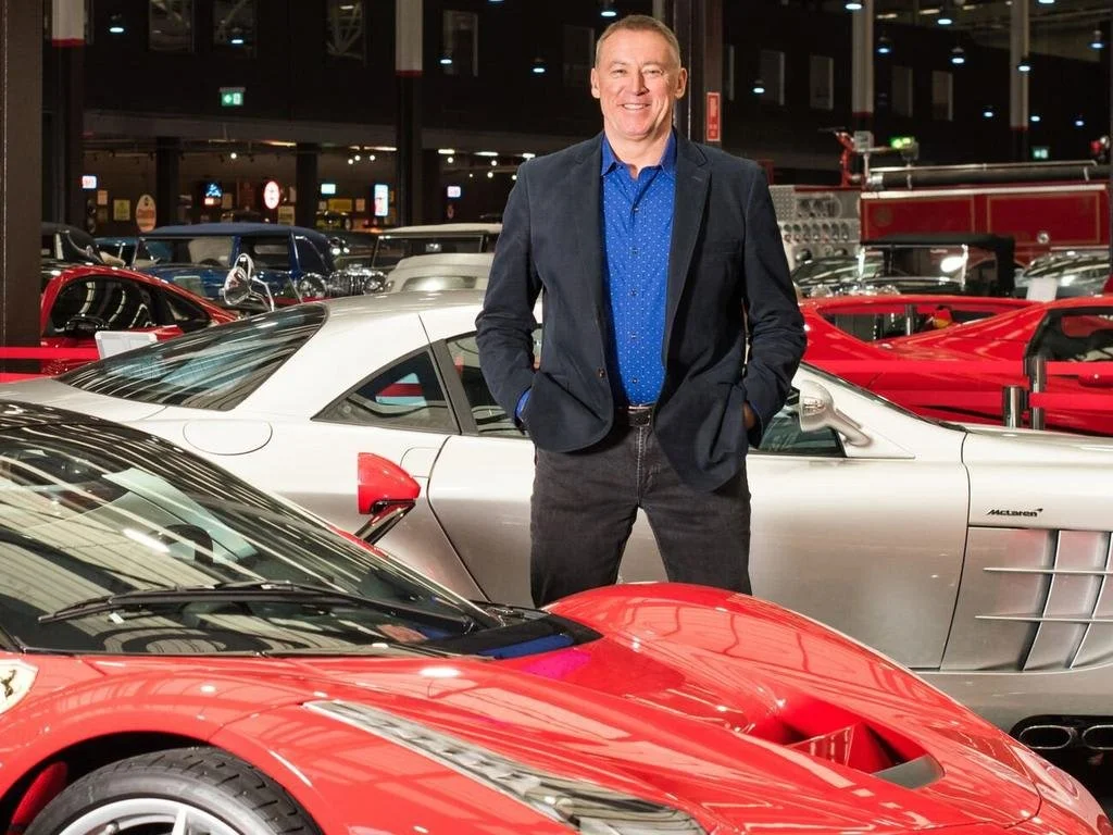 A man in a blue shirt and blazer standing inside a car showroom, surrounded by luxury sports cars including a red car in the foreground and a silver car behind him.