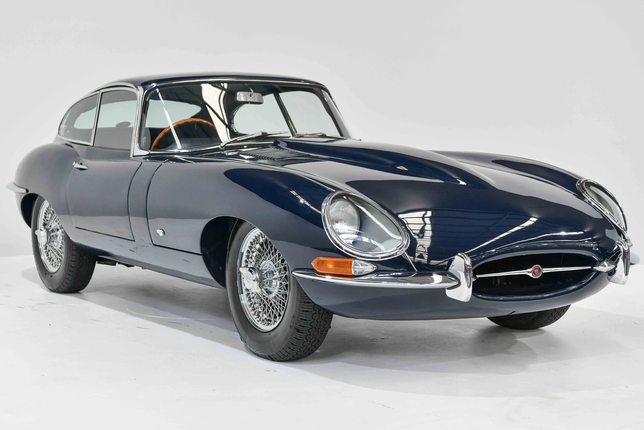 A vintage blue Jaguar E-Type sports car with wire wheels, parked inside a white studio with no other objects around.