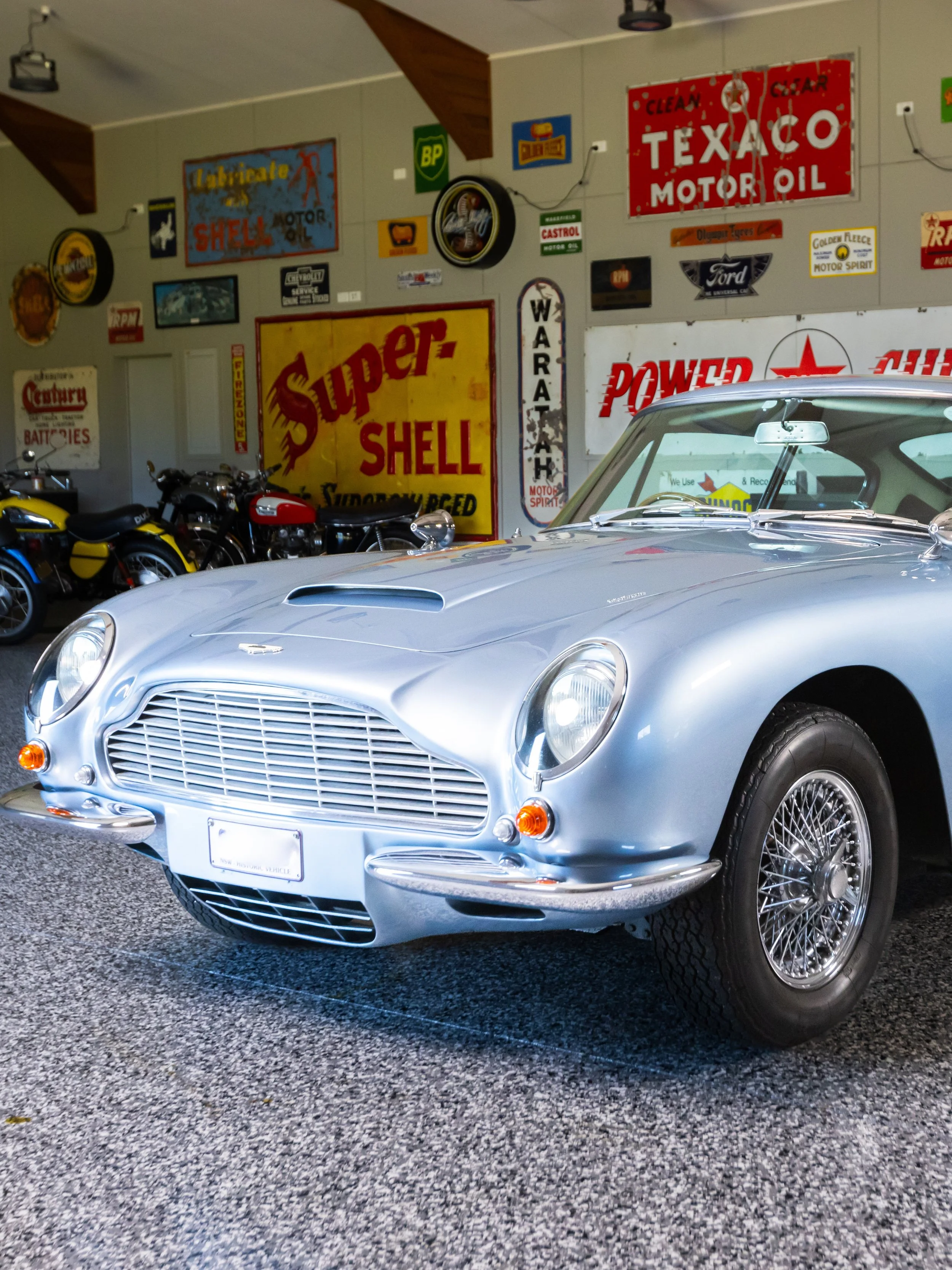 Silver vintage sports car with rounded body and wire-spoke wheels in a garage decorated with vintage automotive signs.