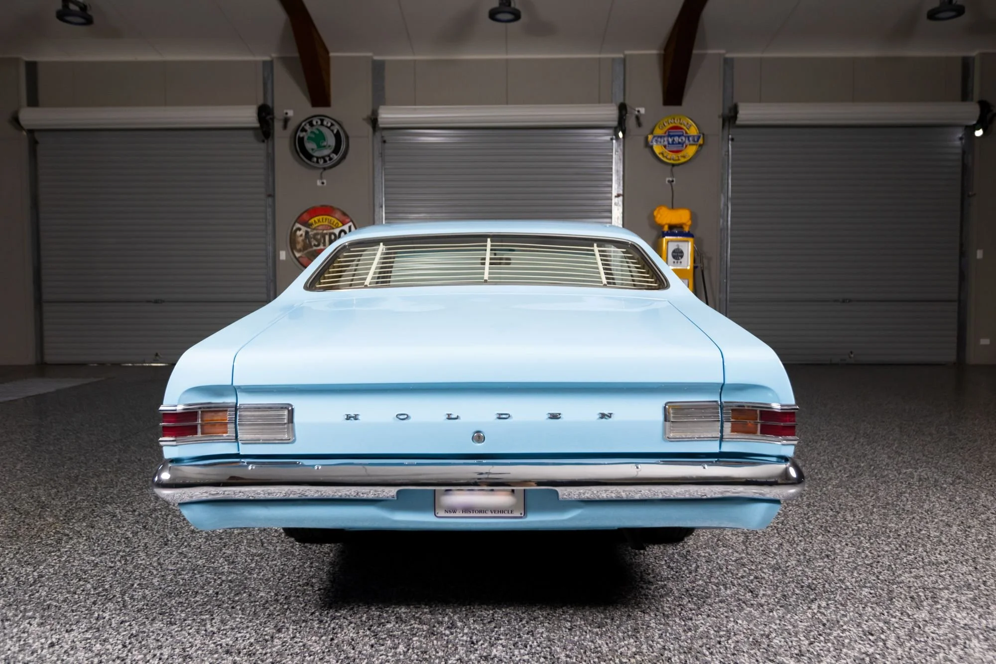 Light blue vintage Holden car in a garage with closed metal shutters, vintage signs on the wall, and a yellow gasoline pump.