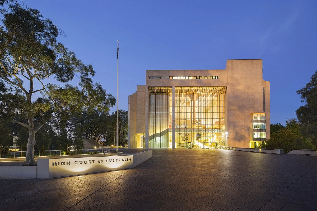 Front view of the High Court of Australia building at dusk, with a large tree on the left, a flagpole, and the building illuminated through large glass windows.