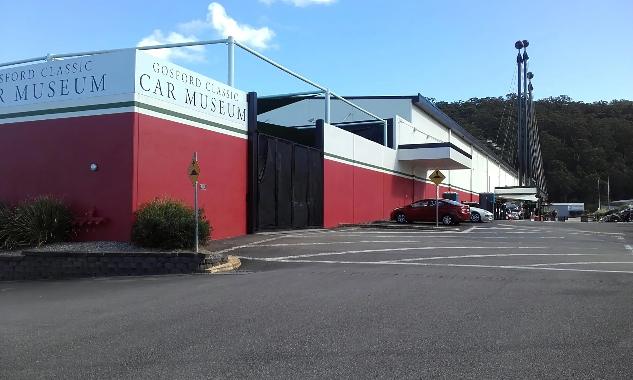 Exterior of the Gosford Classic Car Museum with a parking lot in the foreground and cars parked along the building, which has a red and white color scheme and signs indicating it is a car museum.