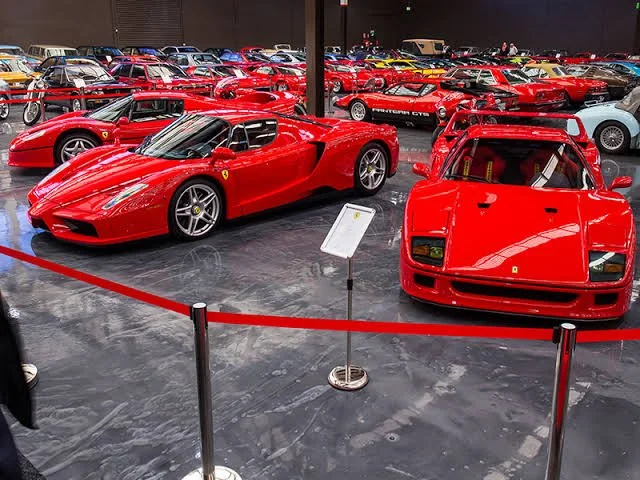 Red Ferrari sports cars displayed in a car showroom, with one modern and one classic Ferrari in the foreground, surrounded by many other Ferrari vehicles in the background.
