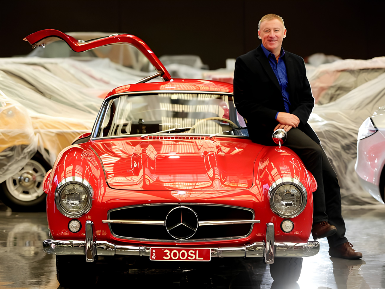 A man in a black blazer and blue shirt sitting on the hood of a red classic Mercedes-Benz 300SL car inside a showroom. The background shows several covered cars.