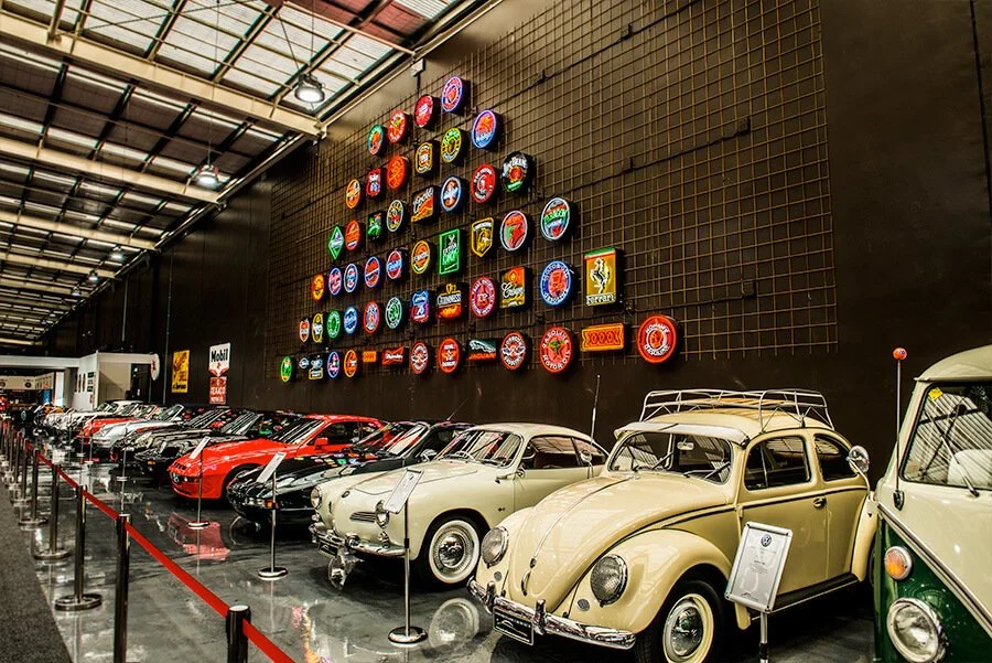 A collection of vintage cars displayed indoors with neon signs on the wall.