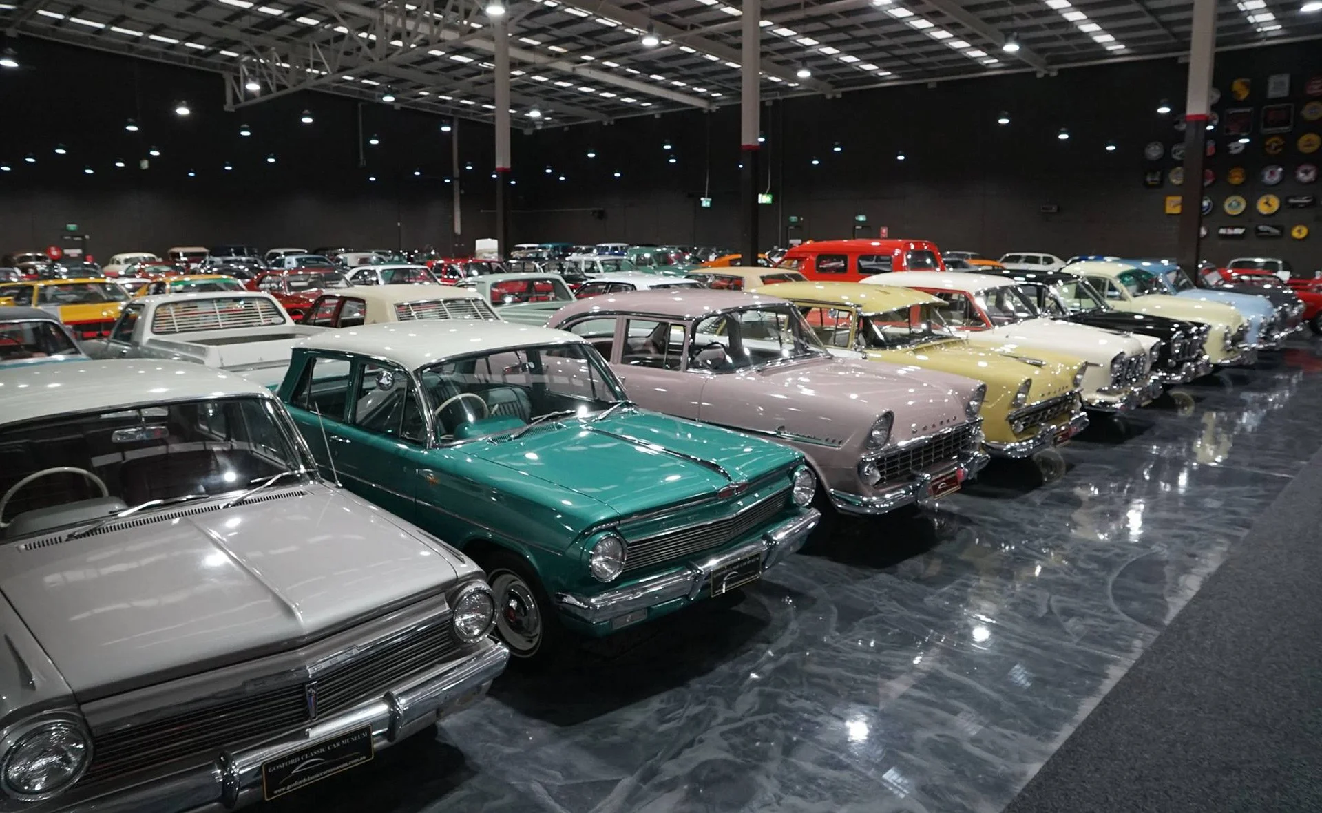 A showroom filled with vintage cars in various colors including green, pink, yellow, white, red, and black, arranged in rows under bright ceiling lights.