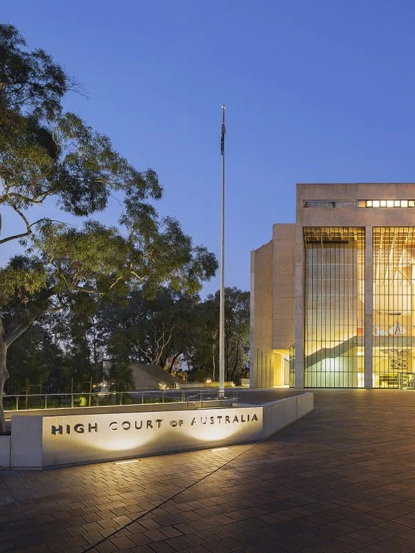 Exterior view of the High Court of Australia building during twilight, featuring a flagpole, the court’s name on a sign, tall trees, and a glass entrance.