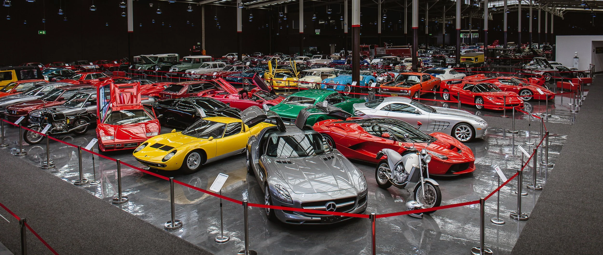 A collection of luxury cars and motorcycles on display in a museum or showroom, including a yellow Lamborghini, a silver Mercedes-Benz, a red Ferrari, and a black motorcycle, all roped off for viewing.